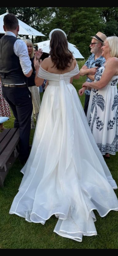 Bride in a flowing white dress, surrounded by wedding guests outdoors.