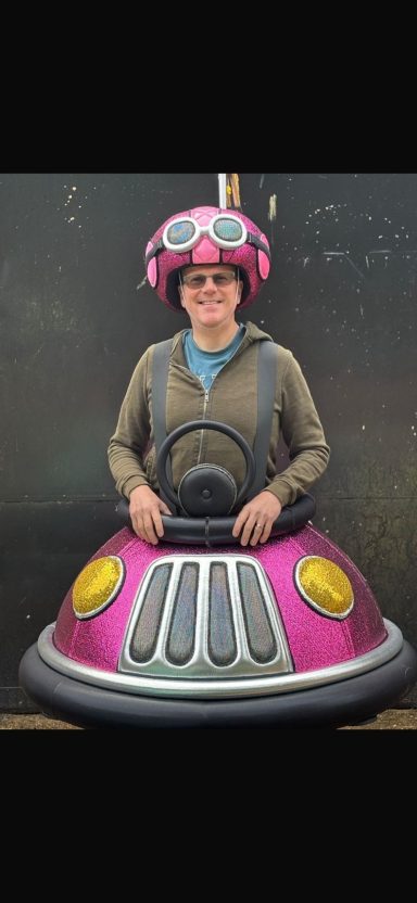 Person in a pink bumper car wearing a pink helmet and glasses, smiling at the camera.