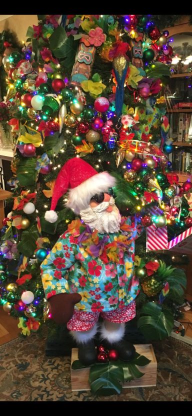 A festive Santa figure in bright floral attire stands beside a colourful Christmas tree.