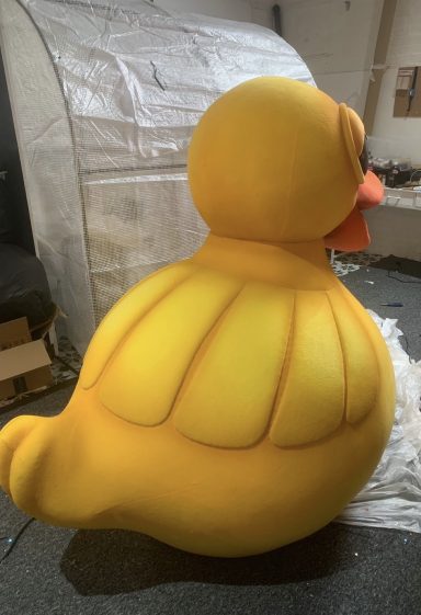 Large yellow rubber duck sculpture, side view, set against a blurred background.