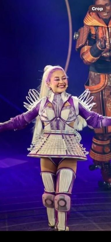 A performer dressed in a purple and white costume, smiling on stage.