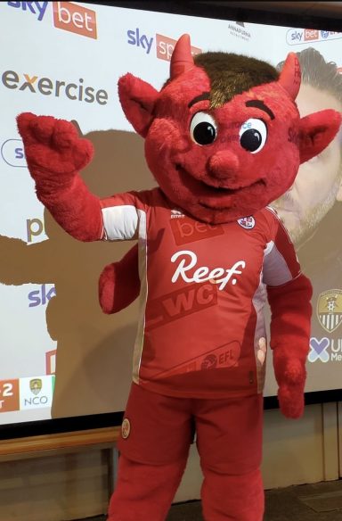 Red devil mascot in a sports jersey, waving cheerfully at an event.