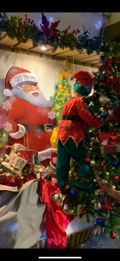 Santa Claus and an elf decorating a Christmas tree with colourful ornaments.