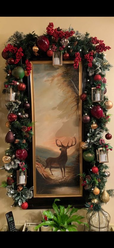 A decorative frame with holly and ornaments surrounding a painting of a deer.