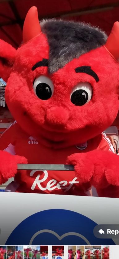 A friendly red mascot with large eyes and a playful expression, holding a sign.