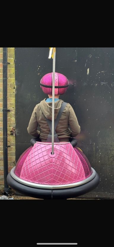 A person sitting in a large pink chair against a wall, wearing a pink hat.