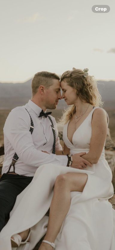 A couple embracing, wearing formal attire, against a scenic outdoor backdrop.
