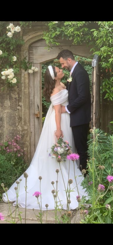 Bride and groom embrace in a garden, surrounded by flowers and greenery.