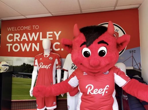 A red devil mascot in a Crawley Town FC kit stands in a club shop.