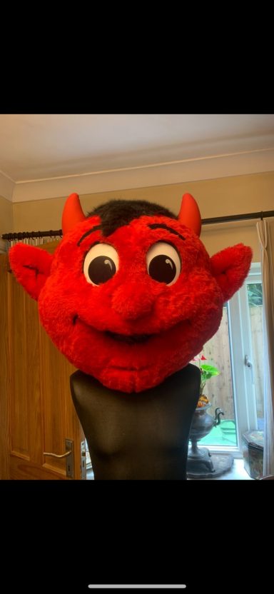 Red cartoon devil's head with big eyes and black horns, worn on a person's hand.