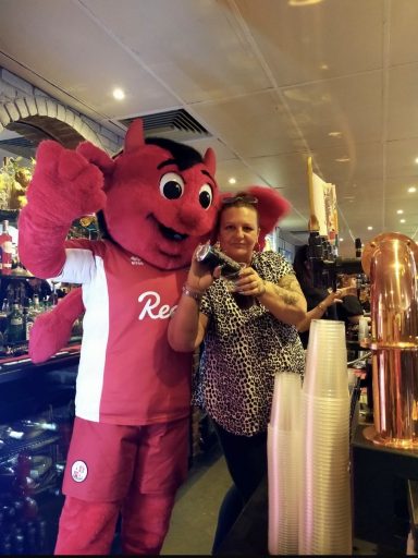 A red mascot in a sports outfit poses with a woman holding a drink in a bar setting.